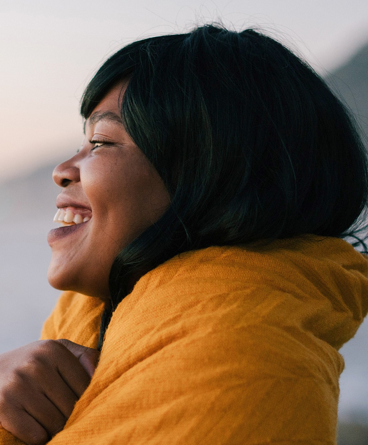 Smiling woman wrapped in yellow sweater outdoors.