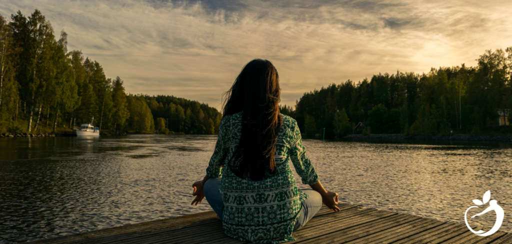 Image of a woman sitting on a dock at sunset meditating, the result of good health coaching.