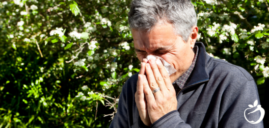 Blog Post Header Image - Managing Allergies. Image of a person standing next to flowers, blowing their nose.
