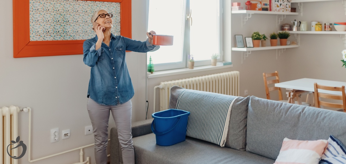 woman on her phone while holding a pan to catch water from a leak in her ceiling